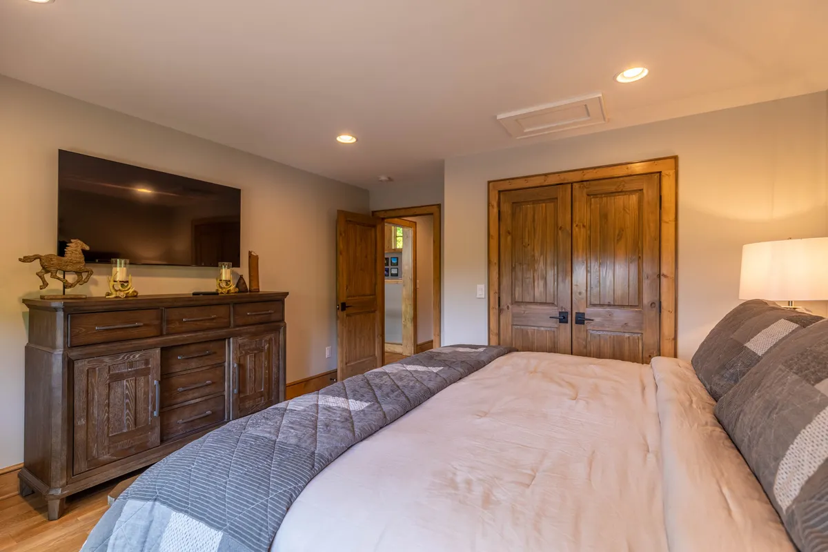 A serene bedroom with a large, inviting bed, wooden furniture, a dresser, wooden doors, and a wall-mounted television. Sunlight streams in, creating a warm and restful atmosphere, ideal for unwinding after a day of adventure.