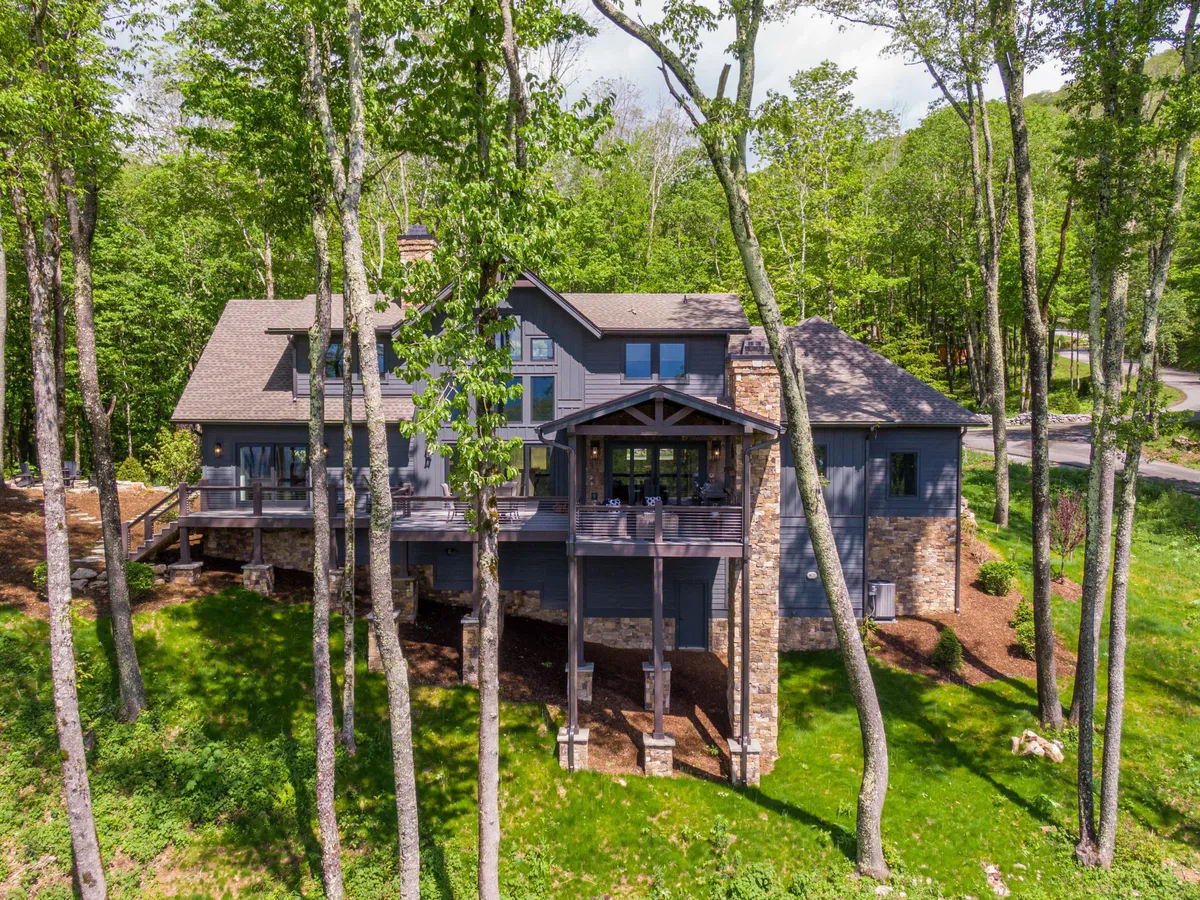 A large wooden deck nestled among tall trees, with the home’s stone facade visible and warm, inviting light spilling from the windows. The surrounding woods create a sense of privacy and connection to nature, while the deck offers ample space for gatherings or relaxation.