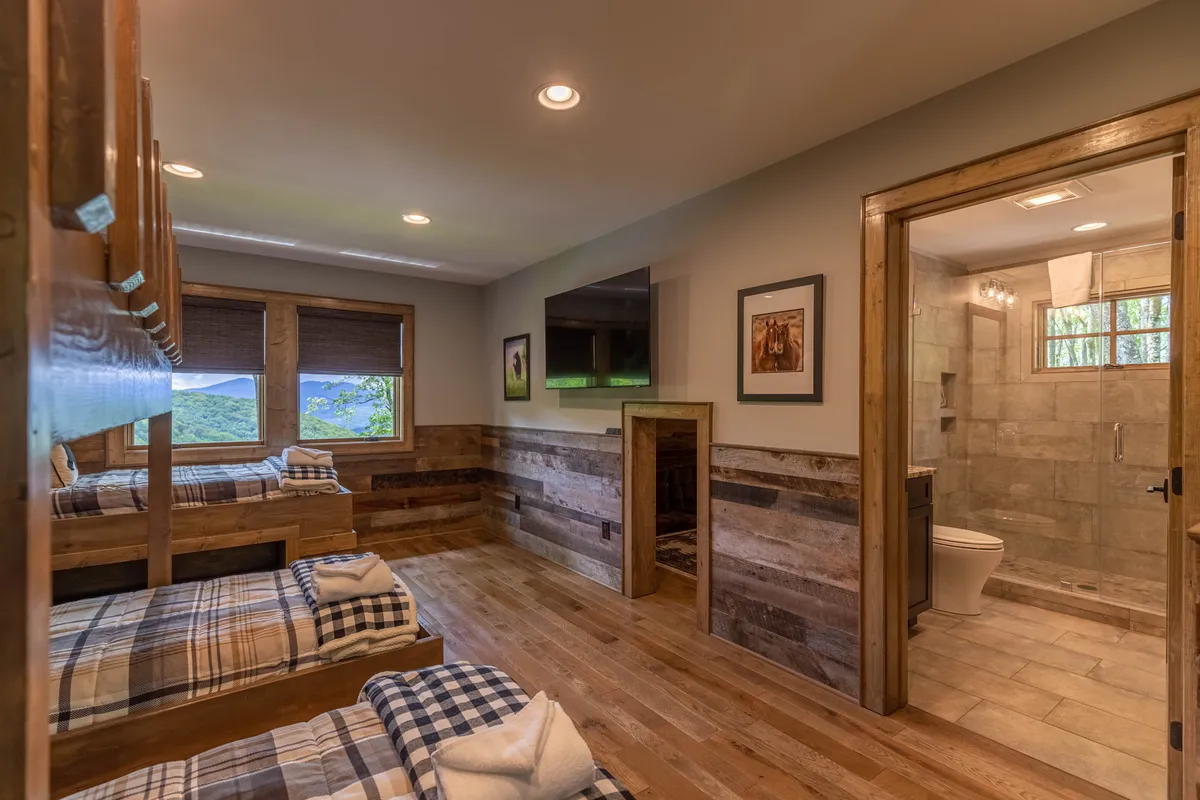 A comfortable bunk room with cozy bunk beds, a window view, framed pictures, a TV, and an entrance to a bathroom. The space is light-filled and designed for both relaxation and fun, ideal for kids and teens.