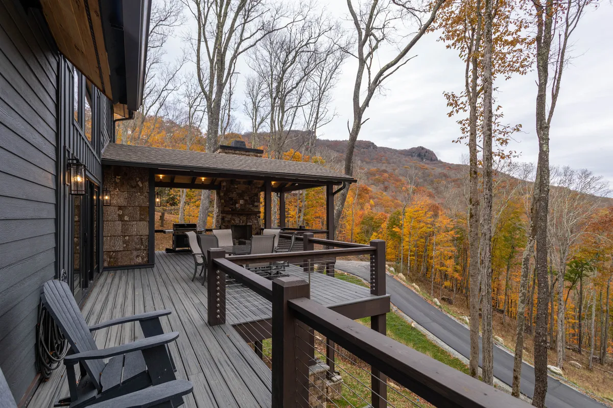 A spacious deck stretches along the house, offering ample outdoor seating with cozy cushions and a BBQ grill. The deck is framed by autumn foliage and overlooks a sweeping mountain view, creating an inviting atmosphere for relaxation or social gatherings.