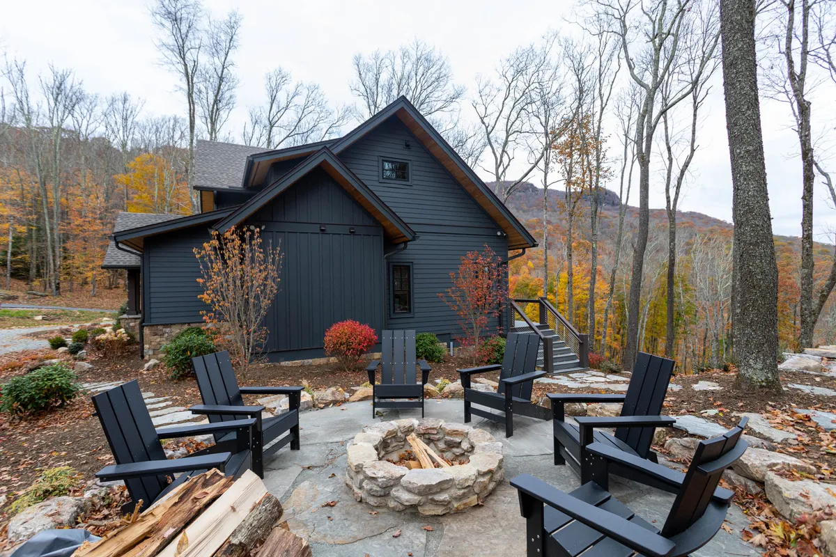 A stone fire pit encircled by Adirondack chairs sits on a natural terrace among mature trees, with the house visible nearby. The setup is both cozy and communal, inviting guests to gather, share stories, and enjoy the outdoors under the stars.