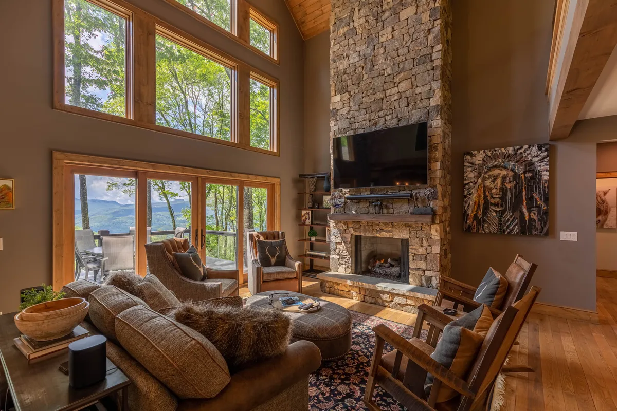 This image showcases an expansive great room featuring a large stone fireplace as its focal point, flanked by comfortable seating and artwork on the wall. Expansive windows flood the space with natural light and offer views of the mountains, creating a dramatic yet welcoming atmosphere. The warm wood tones and plush furnishings invite relaxation and togetherness.