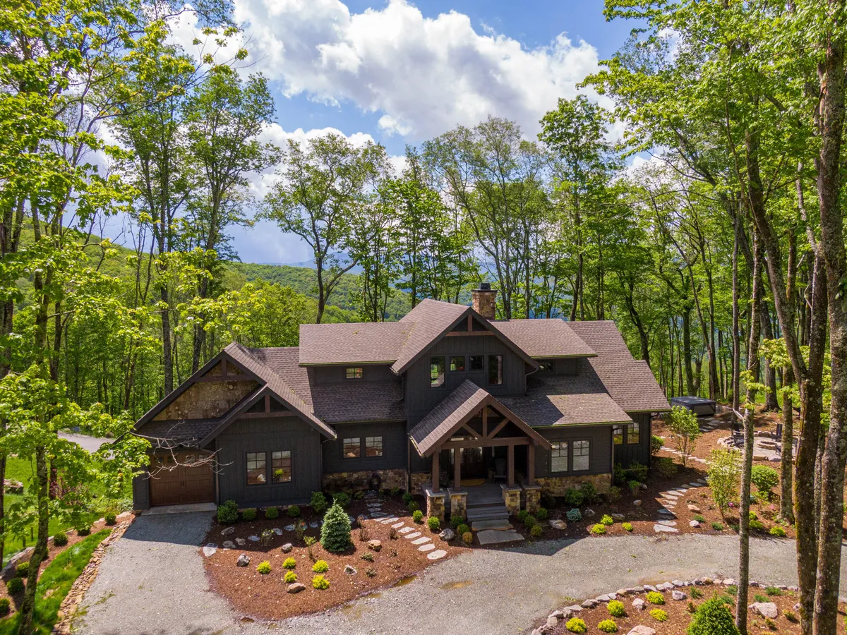 The image showcases the rustic cabin's inviting main entrance, complete with a spacious porch perfect for lounging. Framed by tall trees and featuring a striking stone chimney, the cabin sits harmoniously within the tranquil North Carolina mountain landscape, hinting at sunset views and outdoor relaxation.