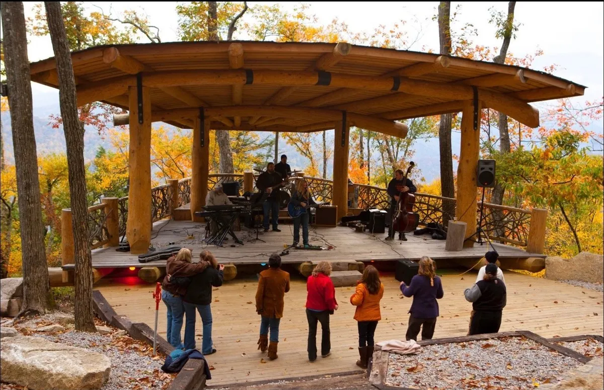 A lively outdoor stage set against autumn foliage, with musicians performing and an engaged audience enjoying the event. The atmosphere is joyful and communal, surrounded by the beauty of the mountains.
