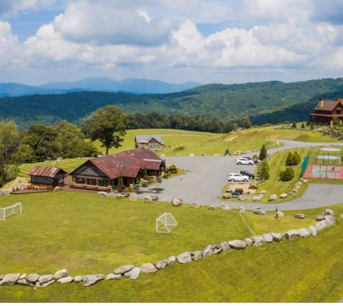An expansive exterior view of a rustic lodge with a red roof, positioned amidst mountains. The scene features visible soccer goals, tennis courts, and open fields, inviting various outdoor sports and family gatherings. The sunset casts a golden glow, enhancing the joyful, communal mood.